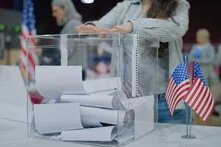 Eine Person wirft einen Stimmzettel in eine durchsichtige Wahlurne. Im Vordergrund stehen zwei kleine US-Flaggen. Weitere Personen sind im Hintergrund.