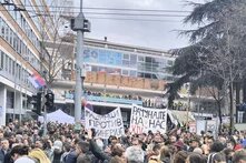 Photo: Protest in Belgrade. A large crowd holds signs with Serbian text. Security forces stand on a staircase in front of a building.