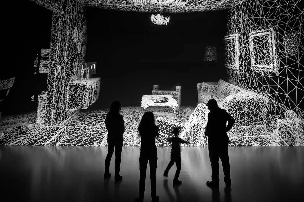 A family of four looks over an art installation that looks like a digitalized version of a living room. Black and white photo